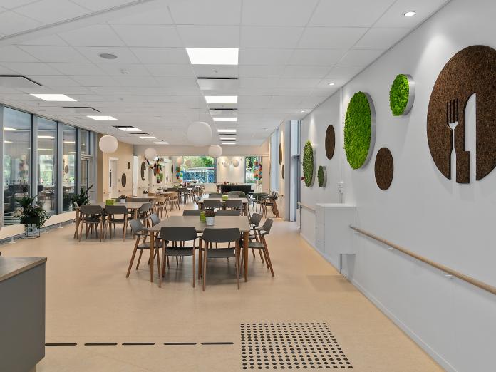 Lyst, aflangt fællesrum med firkantede borde med stole omkring. Den ene væg består af et glasparti og den anden er dekoreret med cirkler af grønne planter og korkplader.