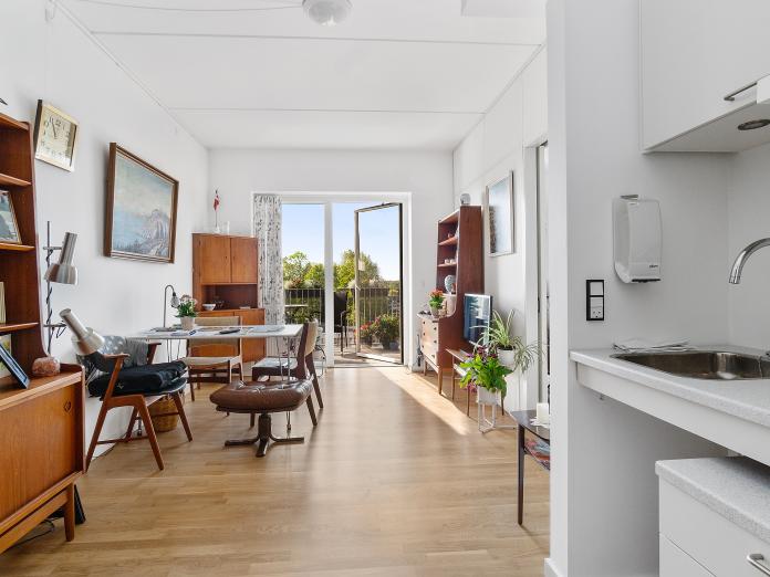 Lyst værelse med stort vinduesparti og dør ud til privat altan. Værelset er indrettet med et tekøkken samt personlige møbler.