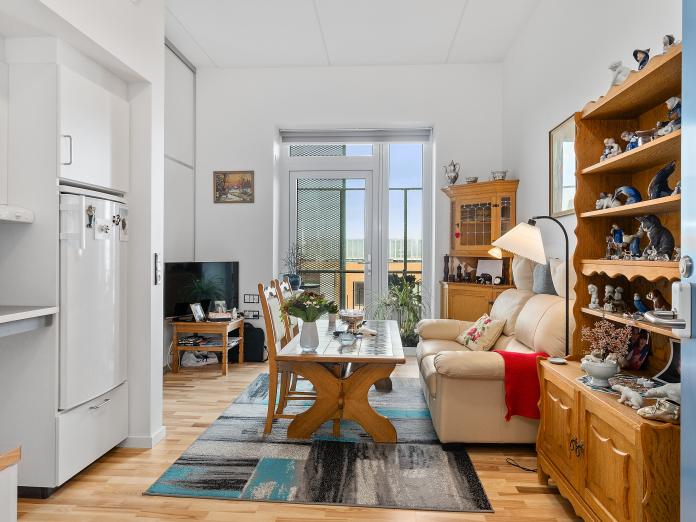 Lys stue med tekøkken og gulv-til-loft-vindue samt dør ud til altan. Stuen er indrettet med personlige møbler som sofa, sofabord, stole og reol.