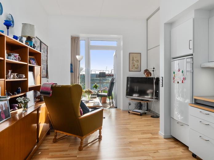 Lyst værelse med tekøkken og åben dør ud til altan. Værelset er indrettet med personlige møbler som en lænestol, et sofabord og et fjernsyn.