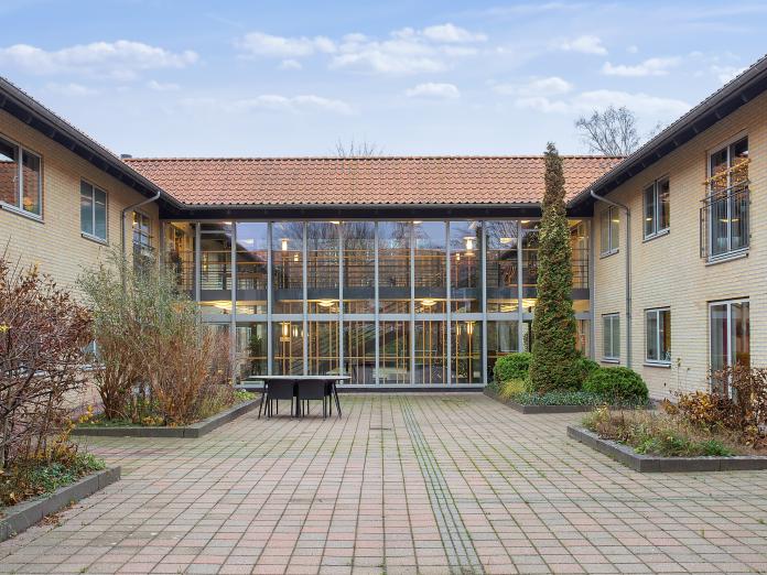 To bygninger i gule mursten forbundet med glaspassage. Foran bygningen er en flisegård bede med buske, træer og blomster.
