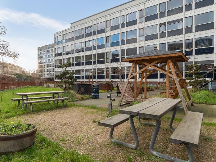 Have med grønt græs, borde-bænke-sæt og dobbelt sofagynge samt planter i krukker, blomsterkummer og træer. I baggrunden et moderne etagebyggeri med store vinduespartier.