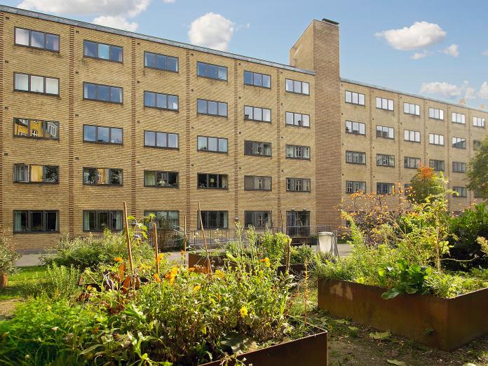Gult murstenbyggeri i flere etager. Foran bygningen er en fliseterrasse og et græsareal med højbede med blomster, urter og grønne planter.