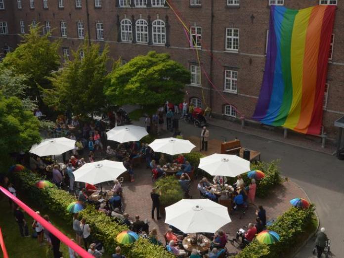 Gårdsplads med mange mennesker siddende omkring runde borde med parasoller og flere mennesker stående. På huset bag gårdspladsen hænger et stort regnbueflag og på en scene står et klaver og en person, der taler.