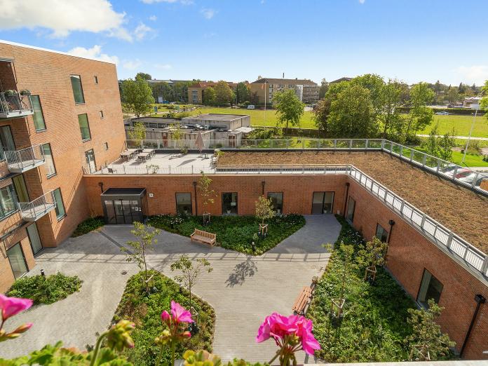 Kig fra privat altan, hvorfra man kan se ned på stor fælles terrasse, et grønt tag og en gårdhave med fliser, bede, træer og bænke. Længere væk ses en grøn park med høje træer. 