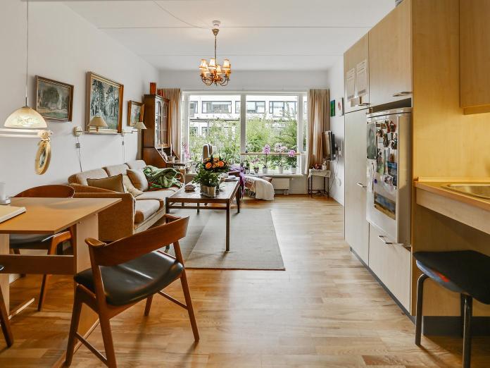 Stue med tekøkken, spisebord og stole, sofa og sofaborde, stort vinduesparti og billeder på væggene.