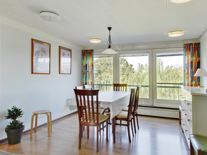 Hyggekrog med stort vinduesparti indrettet med bord med dug og stole omkring samt kommode, billeder på væggen og en grøn plante.