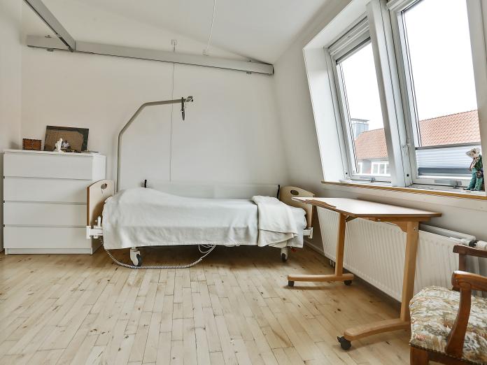 Soveværelse med plejeseng, skråvinduer, lille bord og stol og en kommode. I loftet er der skinner til loftlift.