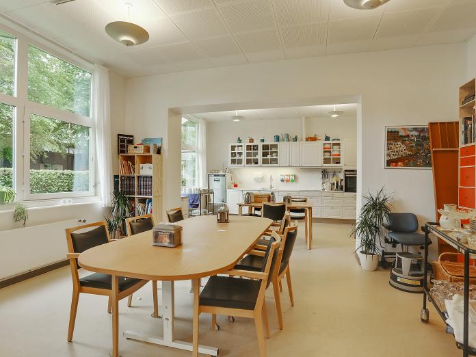 Stort fællesrum med køkken ved den ene væg samt borde med stole omkring og reoler med bøger.