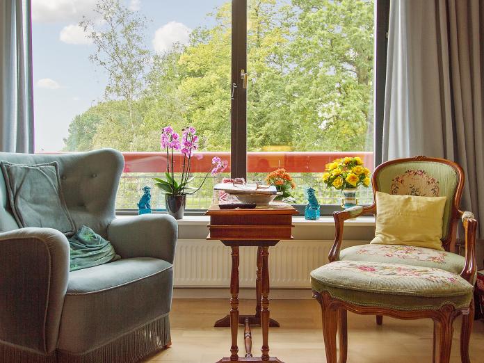 Lænestole foran stort vinduesparti med gardiner og vindueskarme med blomster og personligt nips. 