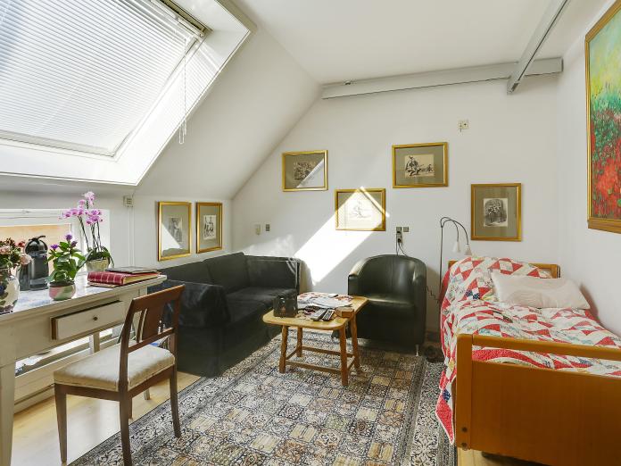Lyst værelse med ovenlysvindue, plejeseng og loftslift. Værelset er møbleret med personlige møbler som sofa, lænestol og sofaborde samt kommoder og et skrivebord med stol. På væggene hænger malerier og billeder og på gulvet er et gulvtæppe.