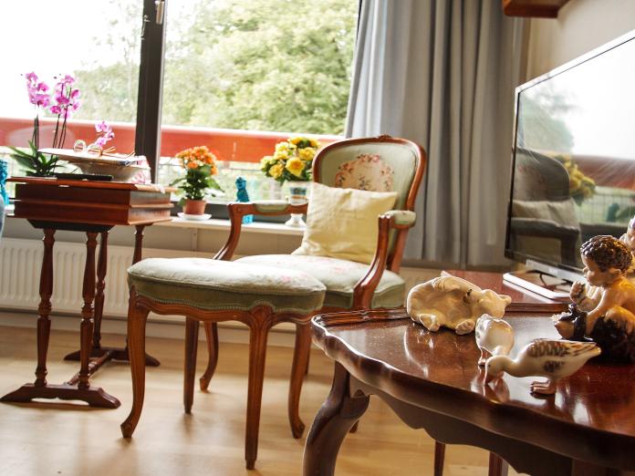 Lænestol og konsolbord foran stort vindue ud mod park. I vindueskarmen står blomster og personligt nips og på et bord står et fjernsyn.