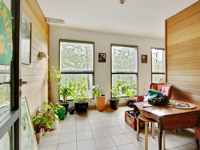 Rum indrettet som terrasse med træbeklædte vægge, fliser på gulvet og masser af grønne planter i krukker og på væggen. Rummet er indrettet med en lænestol og malerier på væggene.