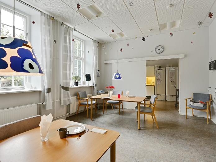 Fælles, lys stue med højt til loftet. Stuen er indrettet med borde og stole og for enden af rummet er der en skydedør ud til et køkken.