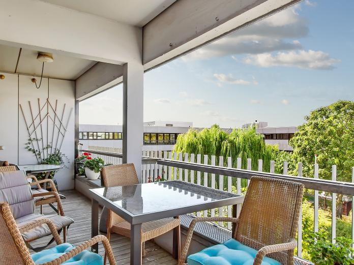 Fælles altan indrettet med bord og kurvestole samt klatregrønt og blomster i krukker. Fra altanen er der kig udover en gårdhave med buske og store træer.
