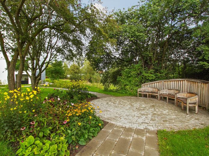 Lille, rund fliseterrasse forbundet med gangstier. Terrassen er indrettet med tre træbænke og omkring terrassen er græs, store træer og bede med forskellige blomster.