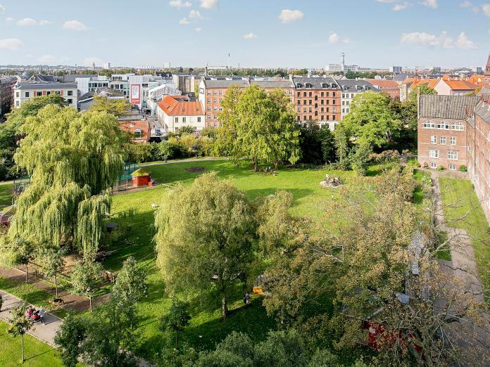 Udsigt højt oppe fra bygning ud over grønt areal med græsplæne, høje træer og legeplads.