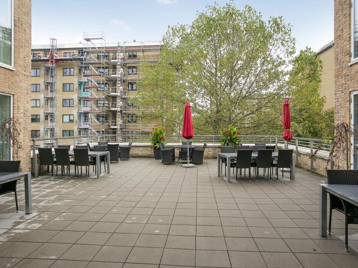 Stor, fælles fliseterrasse med borde og stole samt to røde parasoller og flere krukker med blomster.