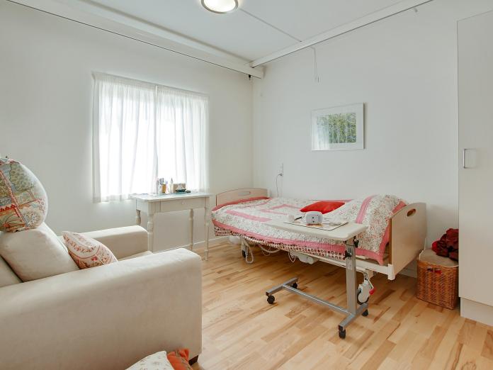 Lyst soveværelse med plejeseng, loftslift og indbygget skab. Værelset er også møbleret med personlige møbler som lænestol og lille skrivebord.