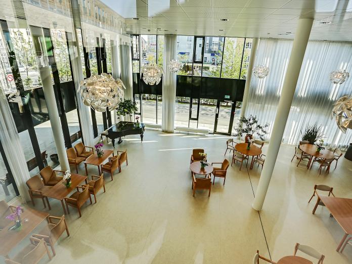 Højloftet fællesrum med store vinduespartier set fra oven. Rummet er indrettet med borde med stole omkring, grønne planter og et flygel. 