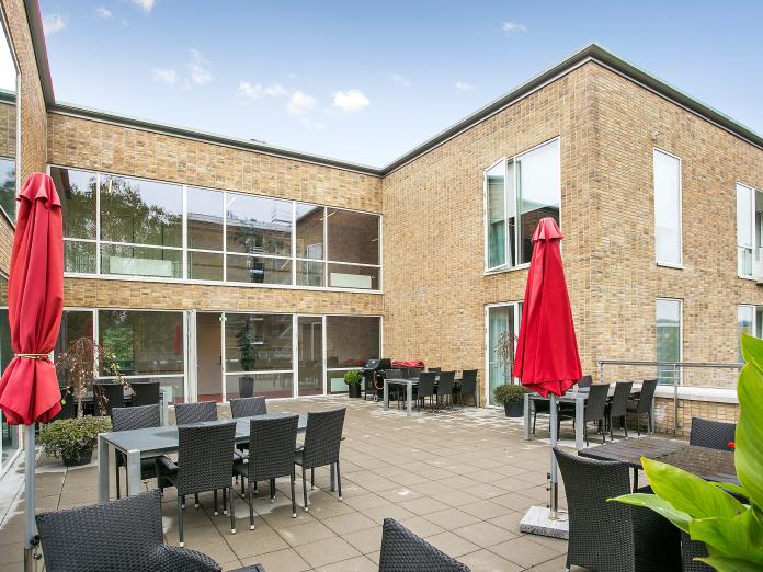 Moderne bygning i lyse sten med store vinduespartier. Foran bygningen er en terrasse med havemøbler, grønne planter i krukker og to røde parasoller. 