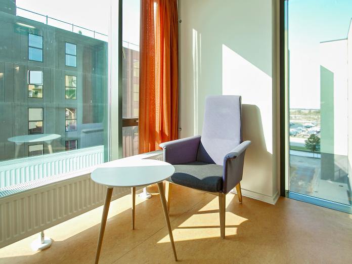 Solrigt hjørne af gangareal med store gulv til loft-vinduer. Hjørnet er indrettet med en lænestol og sidebord.