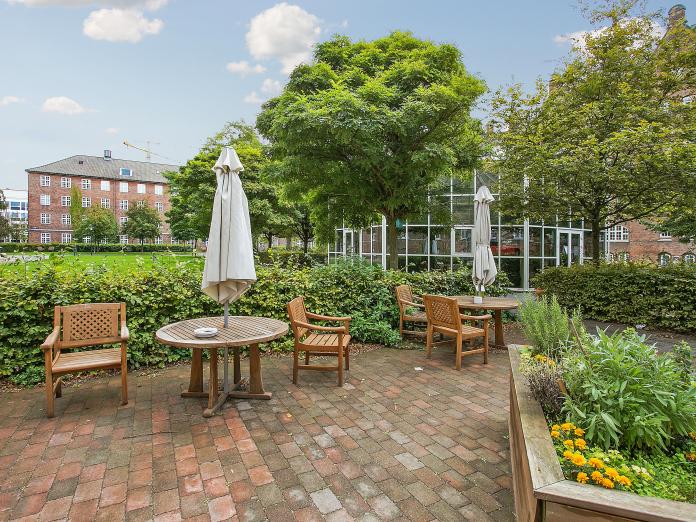 Gårdhave med fliseterrasser indrettet med havemøbler, parasoller og højbede med blomster og krydderurter. Terrasserne er omkranset af hæk og træer og i baggrunden ses et stort drivhus.