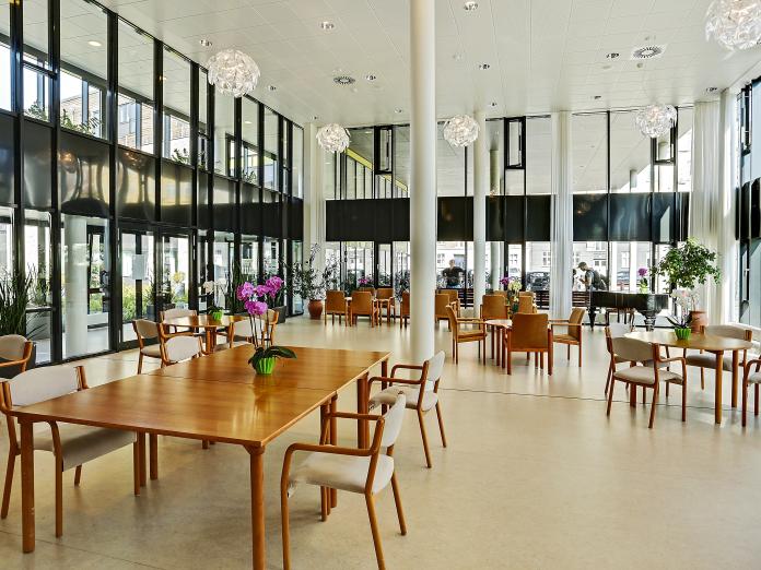 Stort fællesrum med højt til loftet og store glas- og vinduespartier. Rummet er indrettet med borde med stole omkring og på bordene står potteplanter, men der står grønne planter i krukker i hjørnerne.