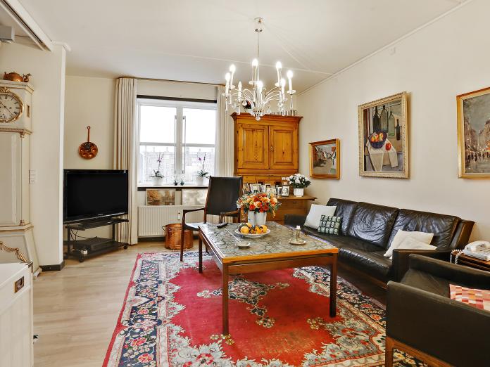 Lys stue indrettet med personlige møbler som sofa, lænestole og sofabord samt chatol, bornholmerur og et fjernsyn. På væggene hænger der billeder og i loftet en lysekrone. I vinduet står to planter.