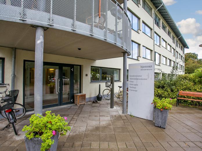 Indgang til Bispebjerghjemmet med et par cykler, en bænk og blomsterkrukker.