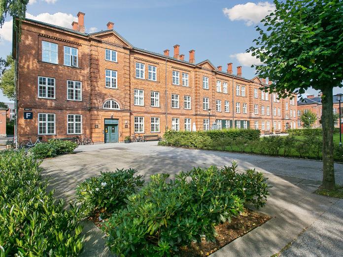 Ældre, rødt firetagers murstensbyggeri med sprossede, hvide vinduer. Foran bygningen er bede med buske omkranset af fliser samt grønt græs omkranset af hæk.