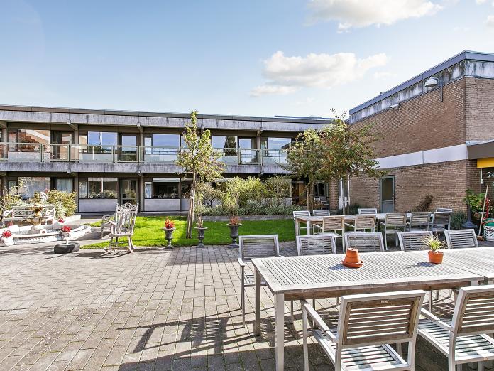 Fliseterrasse med træhavemøbler og blomster i krukker samt grønt græs og træer. I baggrunden er en grå bygning med gule mursten og svalegang.