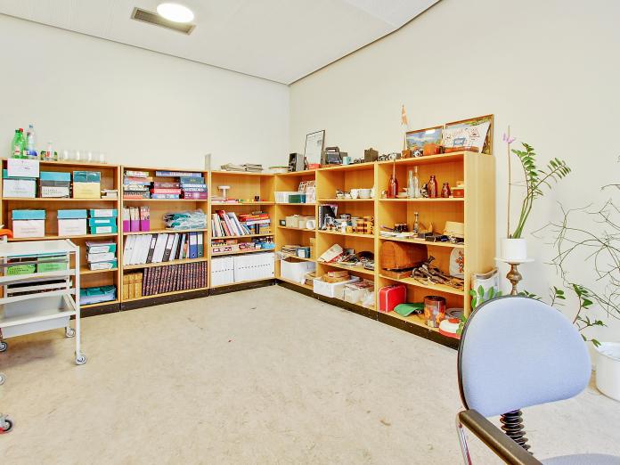 Hvidt rum med reoler med brætspil, bøger, mapper, historiske artefakter samt grønne planter og en kontorstol.