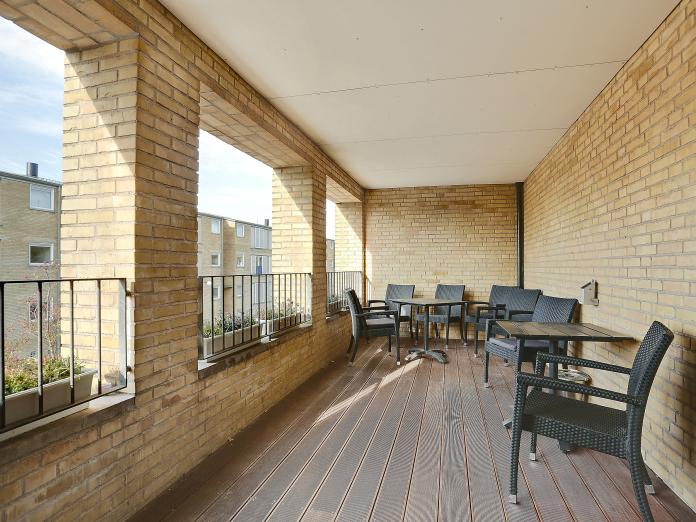 Lukket, fælles altan med gule murstensvægge og trægulv. Altanen er indrettet med sorte altanmøbler samt altankasser med blomster.