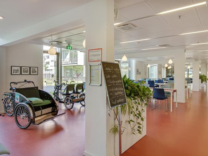 Fællesrum med rødt gulv, hvor der står en ladcykel, en duocykel, en tavle med hvid tekst på, planter og borde i baggrunden.