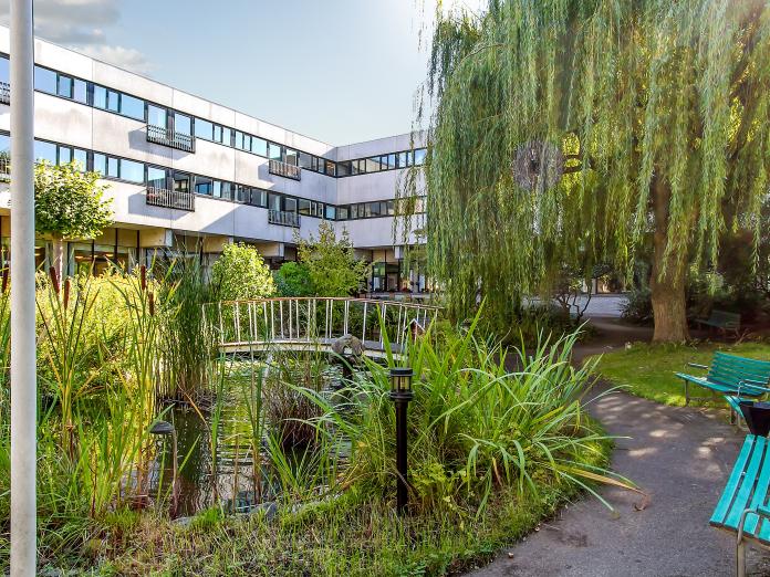 Have med gangstier, bænke og en bro over noget vand. Haven består af træer, grønne planter og græs. I baggrunden ses en moderne bygning med franske altaner.