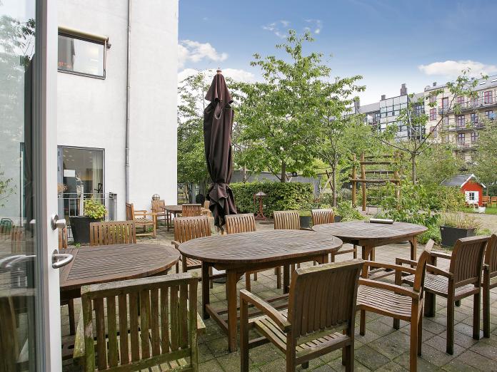 Terrasse med havemøbler i træ, en parasol og kig til gården og mange træer og et lille rødt legehus.