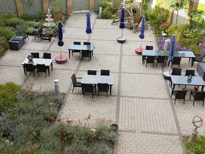 Stor fliseterrasse set fra oven. Terrassen er omkranset af hegn samt buske, bede og træer. Terrassen er indrettet med sorte havemøbler, parasoller og blomster i krukker.