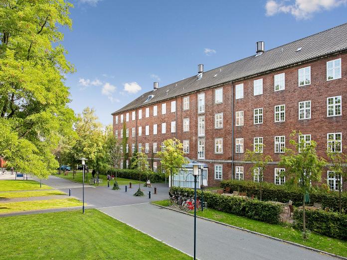 Ældre, rødt firetagers murstensbyggeri med sprossede, hvide vinduer. Foran bygningen er terrasse omkranset af bøgehæk og stor, grøn græsplæne ned høje træer.