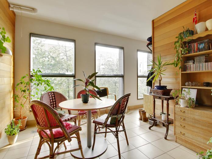 Fællesrum med træbeklædning på væggene og store vinduer. Rummet er indrettet med cafébord og kurvestole samt mange, grønne planter. 