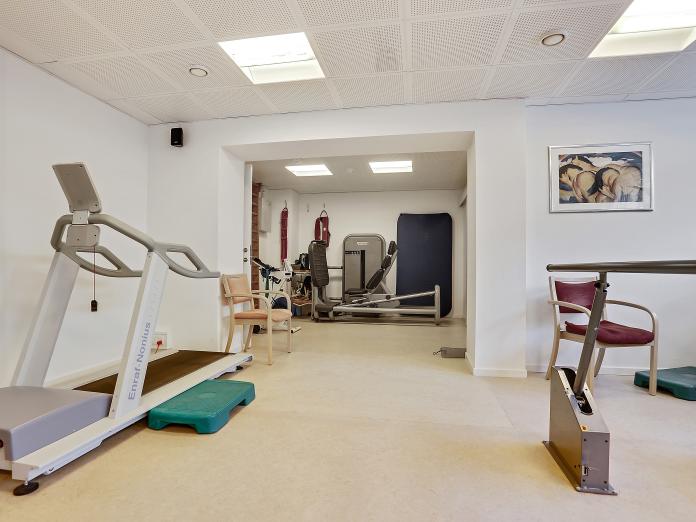 Motionsrum med løbebånd med mulighed for at støtte sig til to armlæn og forskellige maskiner og måtter i baggrunden.