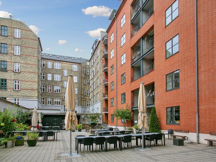 Moderne, rød murstensbygning i flere etager. Bygningen grænser op til andre, ældre bygninger og foran bygningen er en flisegård med havemøbler, bede, buske og grønne planter.