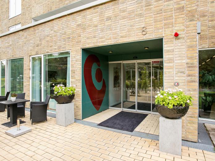 Indgangsparti til bygning i lyse sten med blomsterkummer på søjler og kurvemøbler udenfor.