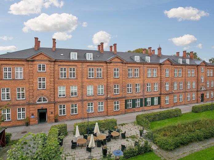 Ældre, rødt murstenbyggeri med hvide vinduer og sort skifertag. Foran bygningen er gårdhaver med fliser og grønt græs omkranset af grønne buske.