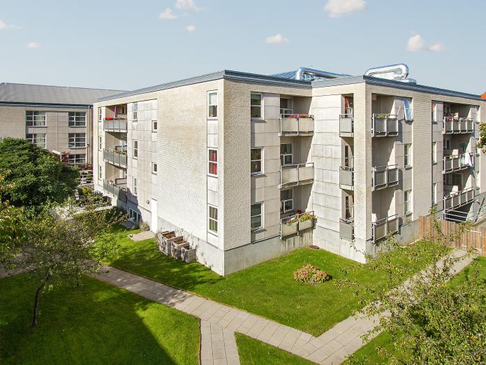 Moderne etagebyggeri med altaner. Foran bygningen er græsarealer med buske, blomster og træer.