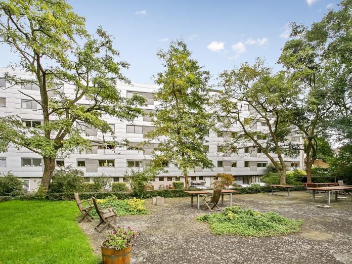 Aftensols grå bygninger set fra terrasse med borde-bænke-sæt, planter, blomster og høje træer.