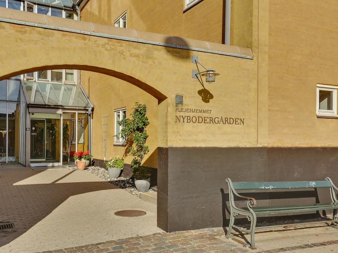 Indgangen til Nybodergården gennem en muret bue ind til selve indgangspartiet i glas. Foran indgangen er en bænk og blomster i krukker.