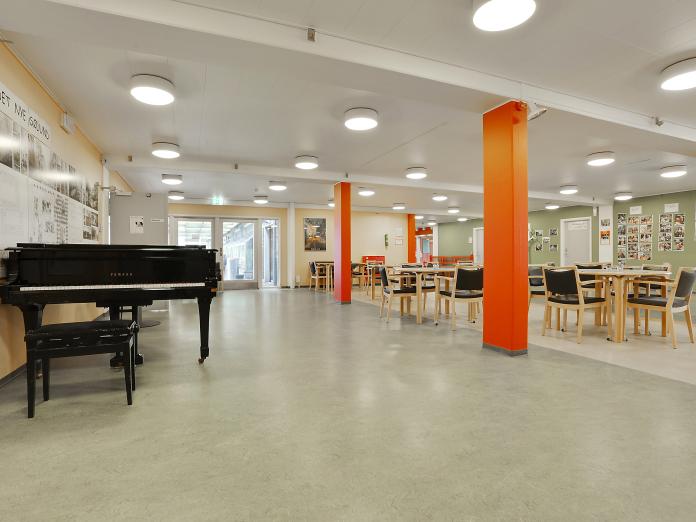 Stort fællesrum med borde og stole samt et flygel og billeder på væggene.