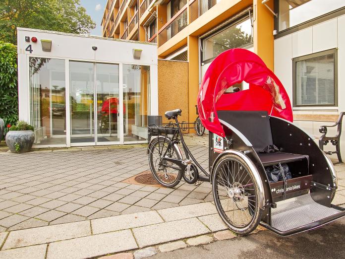 Indgangsparti i glas, som leder ind til en gul bygning. Foran indgangen er krukker med blomster samt en ladcykel indrettet til at transportere personer. 