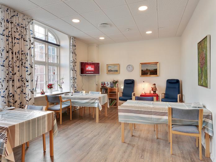 Fællesrum med borde med dug og stole samt et hjørne med lænestole og fjernsyn.